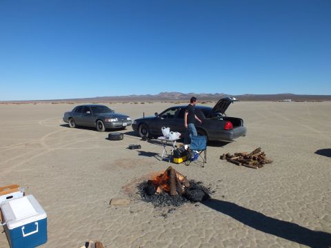2003 Ford Crown Victoria - man works at a sedan's open trunk near a small campfire and cooler; a second sedan and spare tire sit on the lakebed.