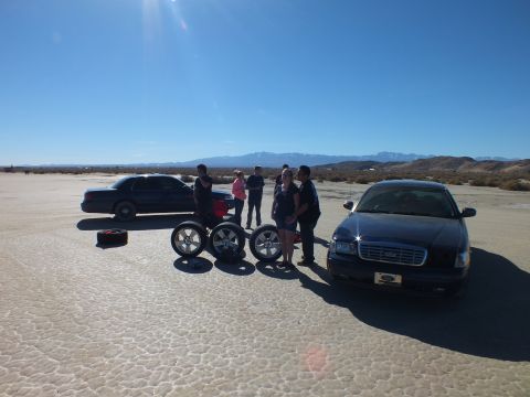 2003 Ford Crown Victoria - People stand between a black Cadillac and another sedan on a desert flat; alloy wheels and tires line up on the ground.