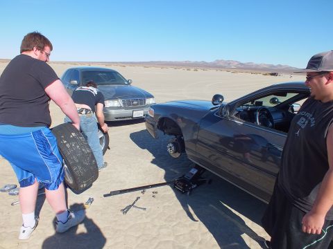 2003 Ford Crown Victoria - Crew swaps tires on a dark coupe next to a Crown Victoria; a floor jack, lug wrench and old tire lie on the sand.