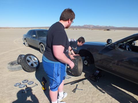 2003 Ford Crown Victoria - man fits a tire to a jacked dark coupe; alloy wheels, hubcaps and tools are spread across the sandy lakebed.
