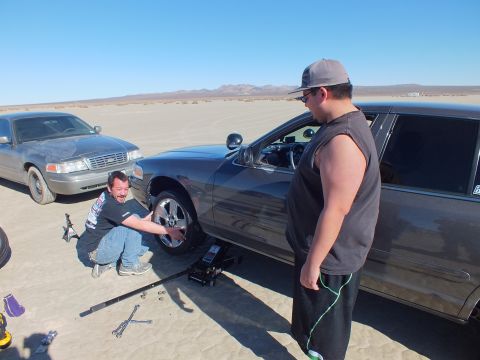 2003 Ford Crown Victoria - One man works the front wheel of a jacked dark sedan as another looks on; a Ford sedan sits in the background.