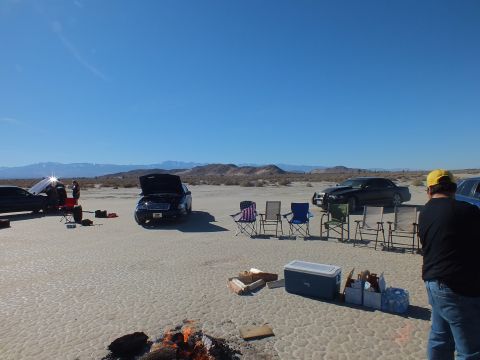 2003 Ford Crown Victoria - man in a yellow cap stands by gear on the lakebed while sedans, an open hood and camp chairs spread across the flat.