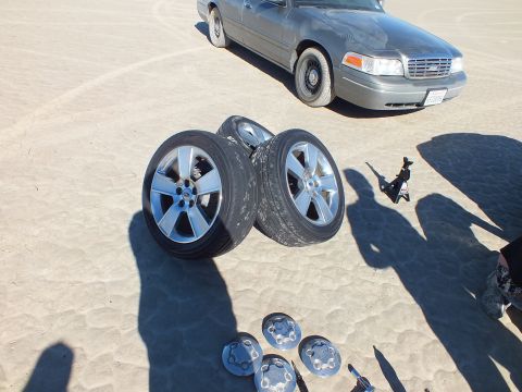 2003 Ford Crown Victoria - Three Ford alloy wheels and removed hubcaps rest on the lakebed sand in front of a grey Crown Victoria sedan.