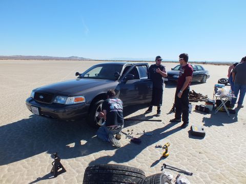2003 Ford Crown Victoria - crew works a jacked grey Crown Victoria on the lakebed; a drill, lug wrench and spare tire lie scattered on the sand.