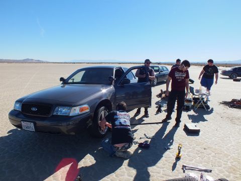 2003 Ford Crown Victoria - Several people change the front wheel of a grey Ford Crown Victoria; tools and a folding chair sit on the lakebed.