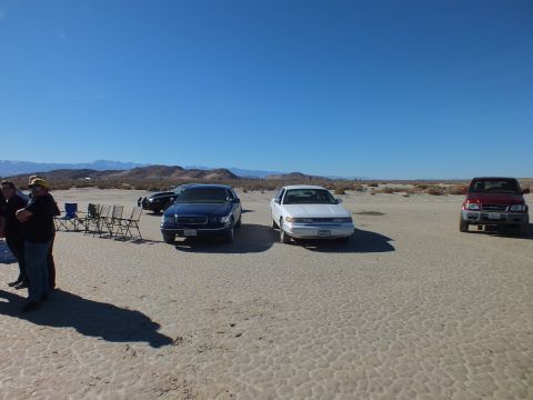 2003 Ford Crown Victoria - Two sedans and a red SUV sit parked on a desert flat; people and empty camp chairs gather at the left edge.