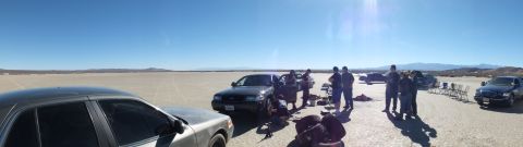 2003 Ford Crown Victoria - Wide panorama of a desert flat: sedans and a group of people spread out beside camp chairs under a clear sky.