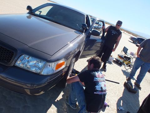 2003 Ford Crown Victoria - person crouches at the wheel of a dark Crown Victoria with its door open while two men stand alongside on the lakebed.