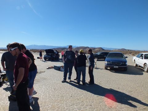 2003 Ford Crown Victoria - group stands talking on the desert flat among parked sedans and a pickup; one car's hood is raised behind them.