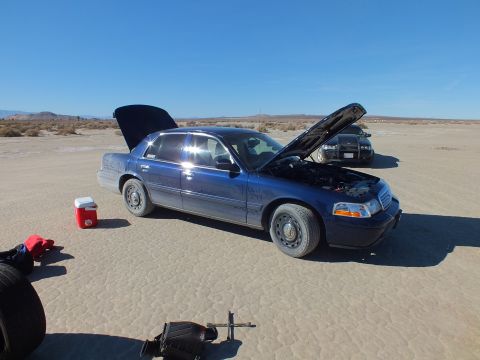 2003 Ford Crown Victoria - blue Crown Victoria sits with hood and trunk open on the lakebed; a tire and lug wrench lie in the foreground.
