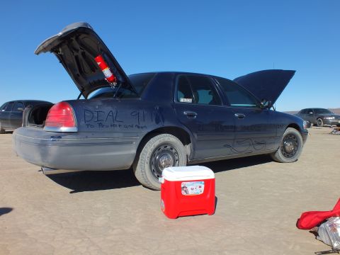 2003 Ford Crown Victoria - dark Crown Victoria sits with trunk open and fire extinguisher mounted; rear marked DIAL 911, red cooler in front.