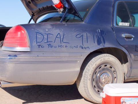2003 Ford Crown Victoria - Rear quarter of a dark sedan scrawled DIAL 911 To Protect and Pawn; steel wheel and a red cooler fill the frame.