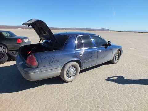 2003 Ford Crown Victoria - Rear-quarter view of a blue Crown Victoria, trunk up, parked on cracked desert flats beside a black sedan.