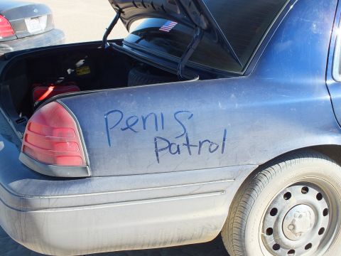 2003 Ford Crown Victoria - Close-up of a dust-coated blue sedan's rear quarter and taillight with crude dust lettering near an open trunk.