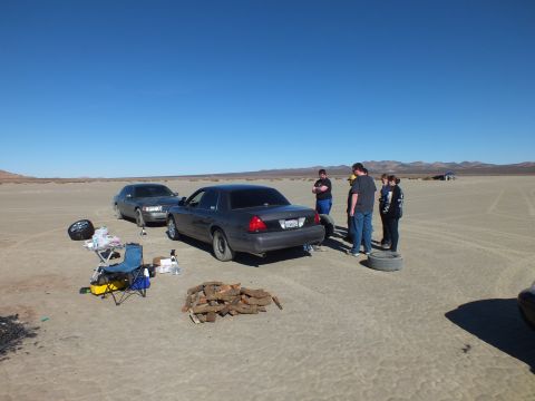 2003 Ford Crown Victoria - Group gathered around two gray sedans with camp chairs, scattered gear and a wood pile on open desert flats.