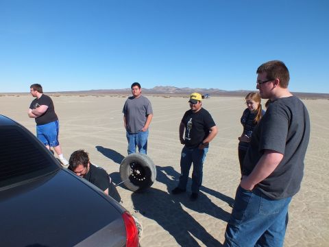 2003 Ford Crown Victoria - Several people gather near a bare steel wheel and a dark sedan on cracked desert flats under clear sky.