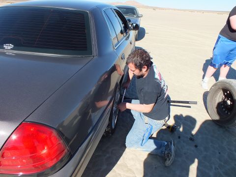 2003 Ford Crown Victoria - man works a tool at the rear wheel of a gray sedan, spare tire nearby, on cracked desert flats.