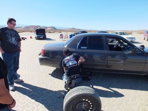 2003 Ford Crown Victoria - Someone works the rear wheel of a black sedan marked INTERCEPTOR while another watches, spare tire in foreground.