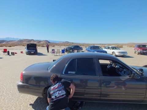 2003 Ford Crown Victoria - person crouches at a black sedan's wheel with more cars and an open hood scattered across the desert flats.