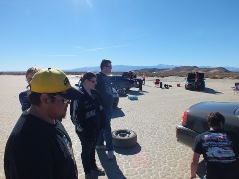 2003 Ford Crown Victoria - Bystanders in cold-weather casual wear watch a tire change near a gray sedan on cracked desert flats.