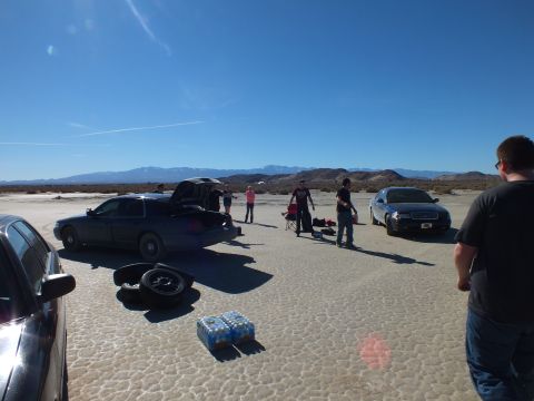 2003 Ford Crown Victoria - Cars parked across cracked desert flats with a case of bottled water, spare tire and people standing about.