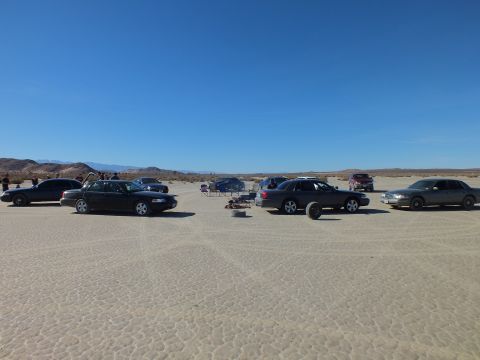 2003 Ford Crown Victoria - Multiple dark sedans spread across an expansive cracked desert flat with distant hills and clear sky.