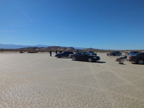 2003 Ford Crown Victoria - Dark sedans and a few people dot a vast cracked desert flat backed by distant brown mountains.