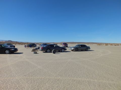 2003 Ford Crown Victoria - cluster of sedans, folding chairs and gear sits on a broad cracked desert flat under clear sky.