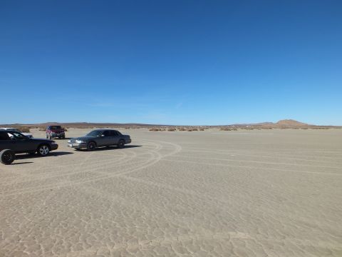 2003 Ford Crown Victoria - few dark vehicles sit at the edge of a cracked desert flat scored with tire tracks near a low hill.