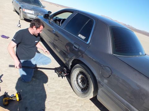 2003 Ford Crown Victoria - kneeling man works a long bar on the jacked rear wheel of a dark Crown Victoria, second sedan behind.