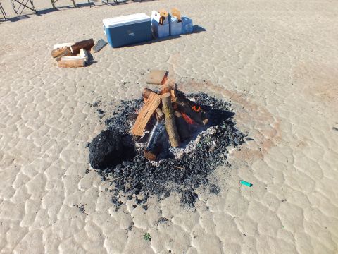2003 Ford Crown Victoria - burned-down campfire of charred logs and ash sits on a cracked desert flat near a blue cooler and firewood.