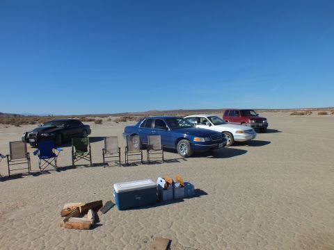 2003 Ford Crown Victoria - blue sedan flanked by a black car and red SUV behind a line of camp chairs, cooler and firewood on desert flats.
