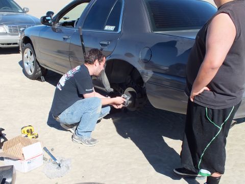 2003 Ford Crown Victoria - man works the rear wheel of a blue sedan with tools and parts box at his feet as another person looks on.