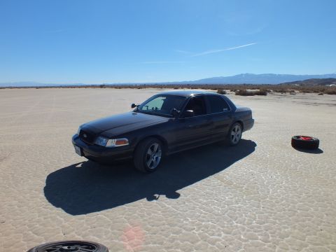 2003 Ford Crown Victoria - lowered black Crown Victoria on alloy wheels sits on a cracked desert flat with spare tires in the foreground.