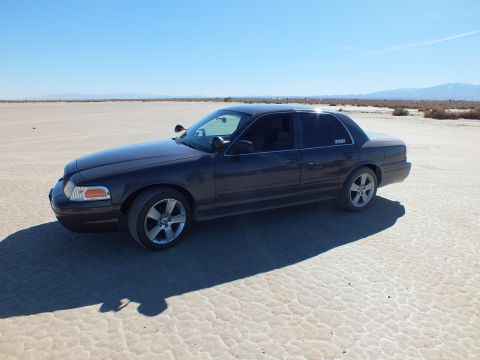 2003 Ford Crown Victoria - Front quarter shot of a dark Crown Victoria on cracked playa under clear blue sky, with distant mountains.