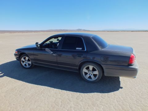 2003 Ford Crown Victoria - Driver-side profile of a dark Crown Victoria on flat playa, alloy wheels and tinted windows visible.