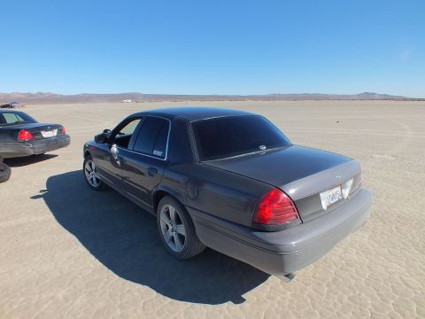 2003 Ford Crown Victoria - Rear quarter of a gray Crown Victoria with red taillights lit, another sedan parked to the left on the lakebed.