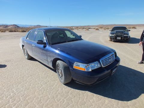 2003 Ford Crown Victoria - Blue Crown Victoria Interceptor with chrome grille parked on playa, a second dark sedan behind it.