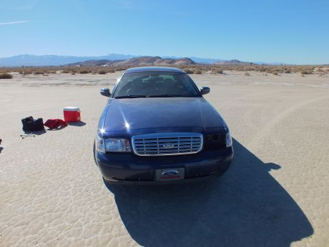 2003 Ford Crown Victoria - Head-on view of a blue Crown Victoria showing chrome grille and Ford badge, gear staged on the playa at left.