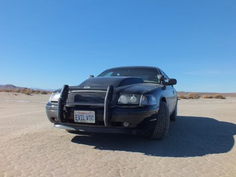 2003 Ford Crown Victoria - Low-angle front quarter of the black EVIL VIC Crown Victoria with chrome guard under deep blue sky.