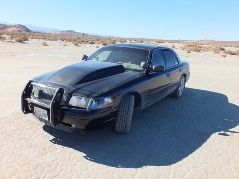 2003 Ford Crown Victoria - Black Crown Victoria with chrome push bar guard on flat lakebed, EVIL VIC plate and desert brush behind.
