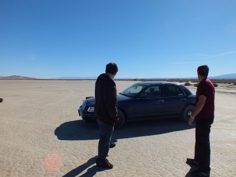 2003 Ford Crown Victoria - Two people stand near a dark blue Crown Victoria on the open playa under clear sky.