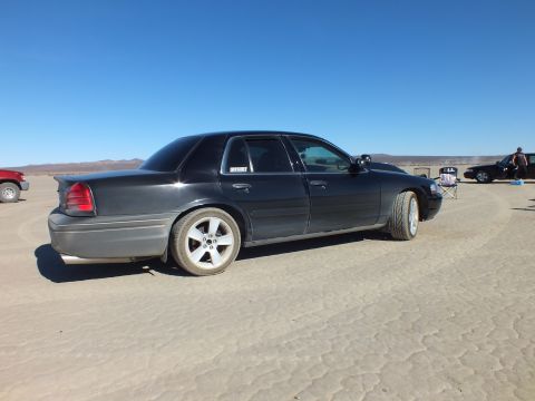 2003 Ford Crown Victoria - Rear quarter of a black Crown Victoria on the playa, alloy wheels and other vehicles in the background.