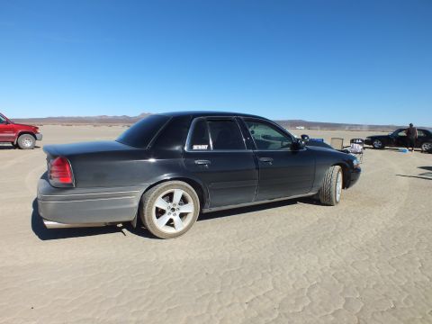 2003 Ford Crown Victoria - Driver-side profile of a black Crown Victoria on the playa, parked vehicles and people in the distance.