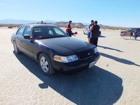 2003 Ford Crown Victoria - Front quarter of a black Crown Victoria with the Ford grille emblem, people and chairs in the background on a lakebed.