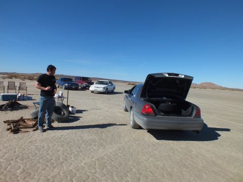 2003 Ford Crown Victoria - Man stands by a gray Crown Victoria with trunk open; spare tires, firewood and other cars sit across the lakebed.