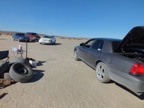 2003 Ford Crown Victoria - Gray sedan with hood and trunk raised next to a folding table and stacked tires on a flat desert lakebed.