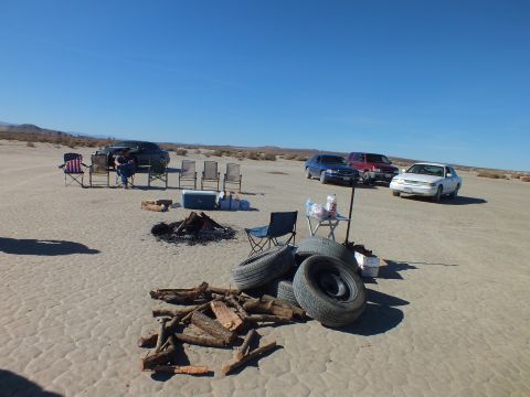 2003 Ford Crown Victoria - Folding chairs, fire pit, stacked tires and firewood arranged near parked vehicles on an open desert lakebed.