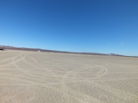 2003 Ford Crown Victoria - Expansive cracked desert lakebed marked with curving tire tracks, low mountains on the far horizon.