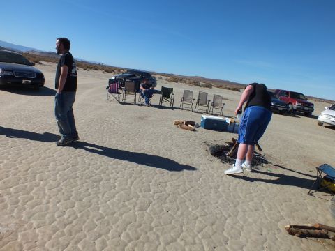 2003 Ford Crown Victoria - Two people tend a fire pit beside a cooler while sedans and an SUV park along the desert lakebed.